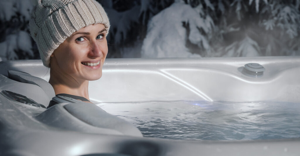 A woman in a winter had sits in a hot tub with snowy trees in the background